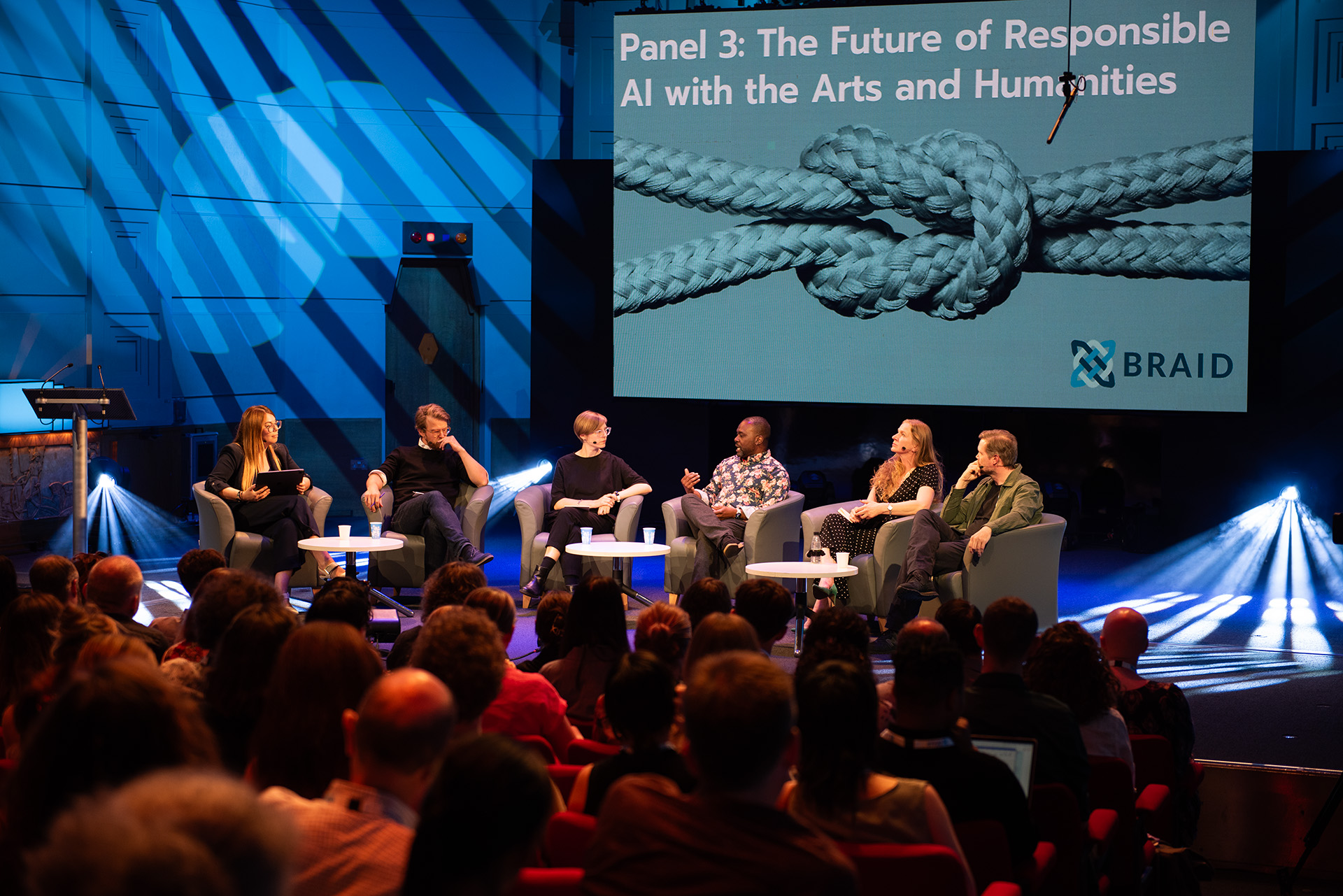 Six speakers seated on stage in chairs, with a screen behind them displaying the panel title, while the audience listen.