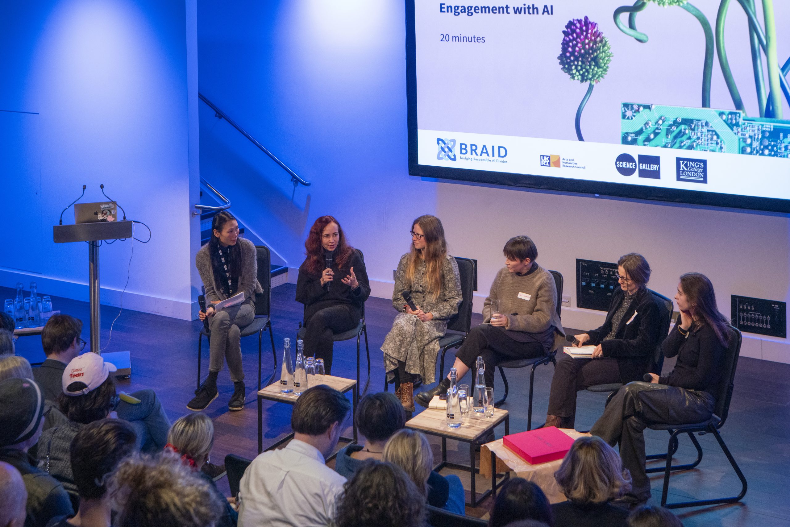 Six people sit on a stage, talking together in front of an audience.