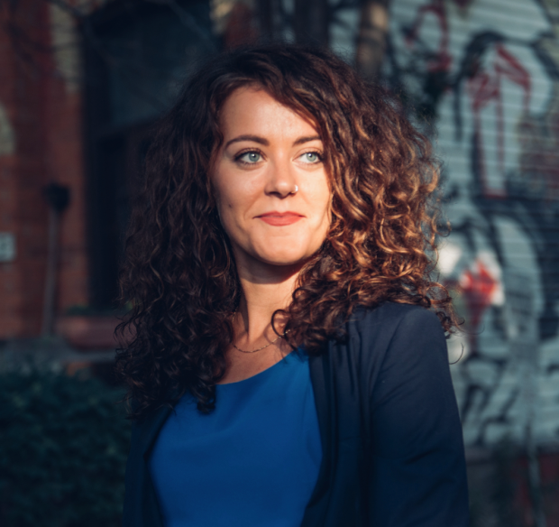 A White woman with brown curly hair, blue eyes, and a blue top smiles to the right of the camera. She is Petra Molnar.