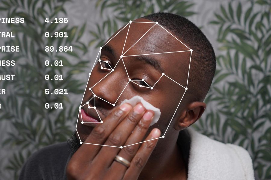 A young black man looks into the camera. He is wearing a dressing gown and has a towel around his neck. He applies a cotton pad to his face and wears a ring on his right hand. a list of words and numbers describe the man’s expression. Happiness 4.185, neutral 0.901, surprise 89.864, sadness 0.01, disgust 0.01, anger 5.021 and fear 0.01.