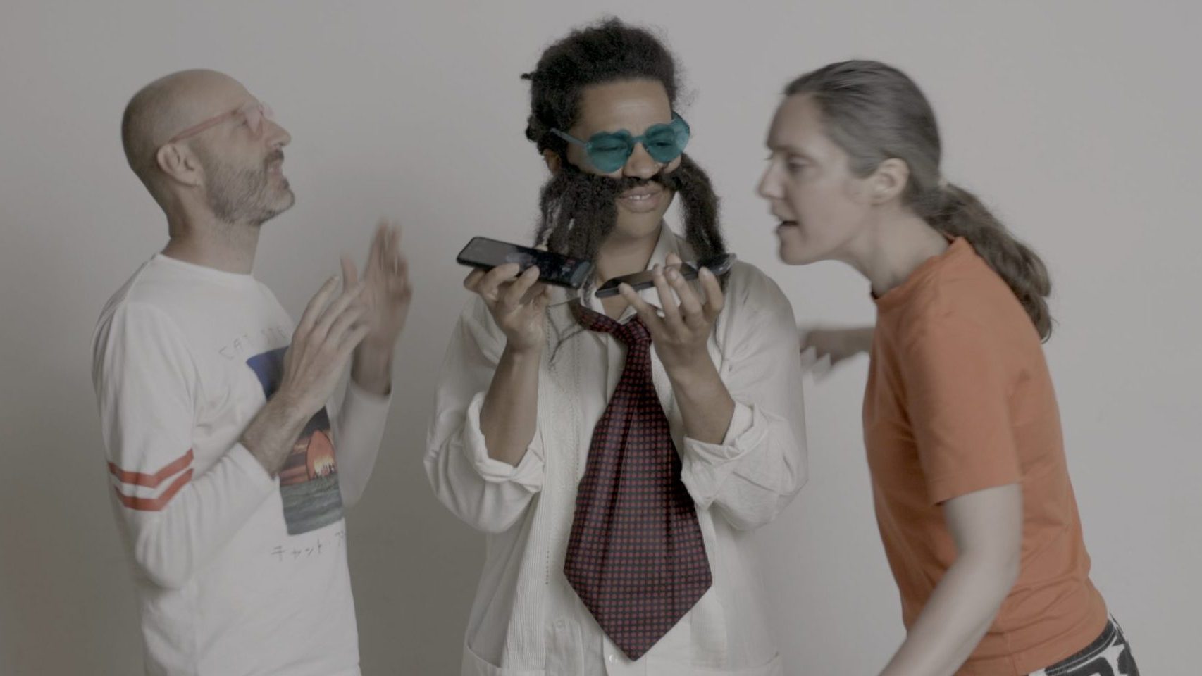 Image shows three people interacting together, playful. One is dressed in clown clothes. They are speaking and listening to two mobile phones at the same time.